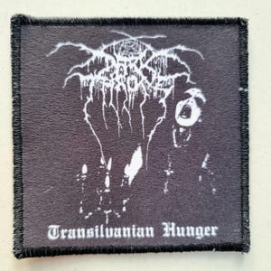 May include: Black square patch with white graphic of a band logo and a screaming figure. The text "Transilvanian Hunger" is printed in white at the bottom. The patch has a black stitched border.