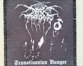 May include: Black square patch featuring a white band logo and a screaming figure graphic. The text "Transilvanian Hunger" is printed in white at the bottom. The patch has a black stitched border.