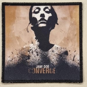 Peut inclure: Écusson carré à bordure noire. Il présente une silhouette d'une personne sur un fond beige. Le texte "JANE DOE CONVERGE" est imprimé en or au bas de l'écusson.