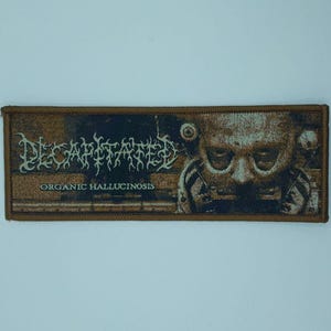 May include: A rectangular patch with a brown border. The patch features the word "DECAPITATED" in a stylized font above the words "ORGANIC HALLUCINOSIS". A detailed illustration of a face with a skull is on the right side.