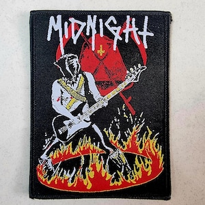 Op de afbeelding: Zwarte en rode geborduurde patch met de bandnaam "MIDNIGHT" in witte letters. De patch toont een figuur die een basgitaar bespeelt voor een rood en wit kruis, omringd door vlammen.