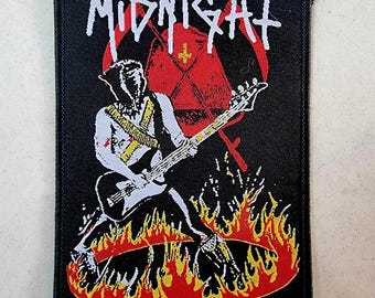 May include: Black and red embroidered patch with the band name "MIDNIGHT" in white lettering. The patch features a figure playing a bass guitar in front of a red and white cross, surrounded by flames.