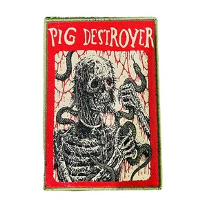 以下が含まれることがあります： 赤い縁取りと白い文字で「PIG DESTROYER」と書かれた長方形のパッチ。パッチには、ヘビと赤い目を持つ骸骨の姿の白黒イラストが描かれています。