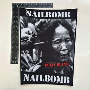 May include: Black and white patch featuring the word "NAILBOMB" above and below a grayscale image of a person. The text "POINT BLANK" is in a red rectangle. The patch is rectangular.