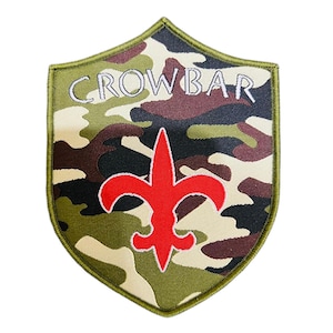May include: Shield-shaped patch with a camouflage pattern in shades of green, brown, and beige. The word "CROWBAR" is embroidered in white across the top. A red fleur-de-lis is centered below the text. The patch has a green border.