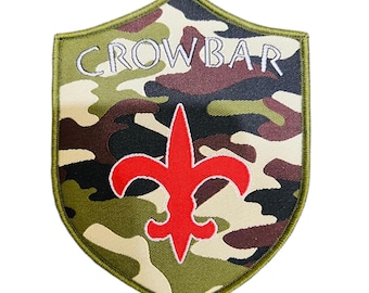 May include: Shield-shaped patch with a camouflage pattern in shades of green, brown, and beige. The word "CROWBAR" is embroidered in white across the top. A red fleur-de-lis is centered below the text. The patch has a green border.