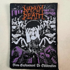 May include: Black and white patch with the band name "Napalm Death" in orange letters. The patch features a graphic of a person with their hands over their face, surrounded by other figures. The text "From Enslavement To Obliteration" is at the bottom of the patch.