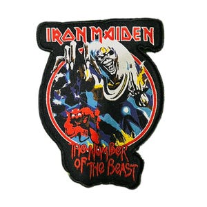 Könnte beinhalten: Gestickter Aufnäher mit dem Iron Maiden-Logo in Rot. Der Aufnäher zeigt den Text "Iron Maiden" über einem kreisförmigen Bild einer skelettartigen Figur mit weißen Haaren. Der Text "The Number of the Beast" ist ebenfalls vorhanden.
