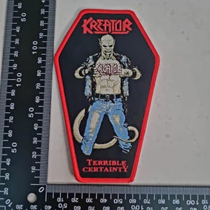 Könnte beinhalten: Schwarzer Aufnäher mit rotem Rand, mit dem Bandnamen "Kreator" in Rot. Der Aufnäher zeigt eine muskulöse Figur in blauen Jeans und Lederjacke, mit dem Wort "Kreator" auf der Brust. Der Text "Terrible Certainty" steht unten.