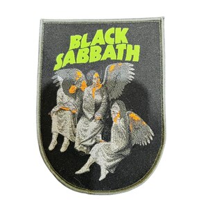 Puede incluir: Parche bordado con la inscripción "BLACK SABBATH" en verde neón. Muestra tres figuras con alas de ángel en túnicas grises con detalles naranjas, sobre un fondo negro.
