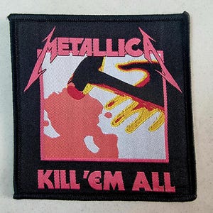 May include: Black fabric patch with the band name "Metallica" in pink lettering and the album title "Kill 'Em All" in pink lettering. The album cover art features a red and white hand with a black arm reaching down.