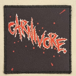 May include: Square black patch with the word "CARNIVORE" in jagged red lettering. The patch has a black border and small red splatters. The design is reminiscent of heavy metal band merchandise.