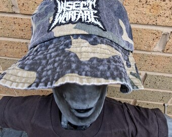 May include: Camouflage bucket hat with the text "INSECT WARFARE" in a black patch. The hat features a green, brown, and grey pattern. The hat is worn by a statue.