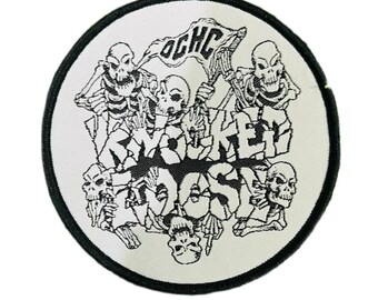May include: Circular patch with a black border. The design features a white background with a black and white graphic of skeletons and the words "ROCKET OOSH". A flag with the letters "OGHC" is also included.