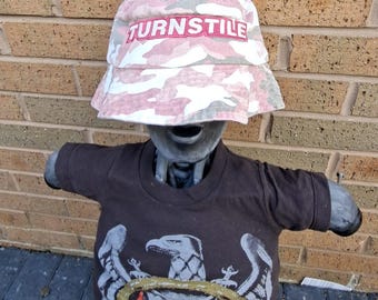 May include: A mannequin wearing a pink and white camouflage bucket hat with the word "TURNSTILE" in pink lettering. The mannequin also wears a dark brown t-shirt with an eagle graphic and the word "SLAYER" in red.
