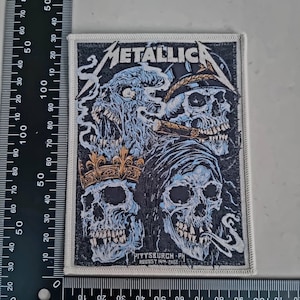 Peut inclure: Écusson rectangulaire avec un motif de crâne et le mot "METALLICA". L'écusson a un fond noir avec des crânes bleus et blancs, et le texte "PITTSBURGH, PH AUGUST 14TH, 2022". Mesure environ 9 cm sur 7,6 cm.