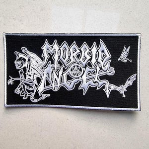 May include: A black embroidered patch with white stitching. The patch features the text "MORBID ANGEL" in a gothic font with a winged demon figure and a pentagram.