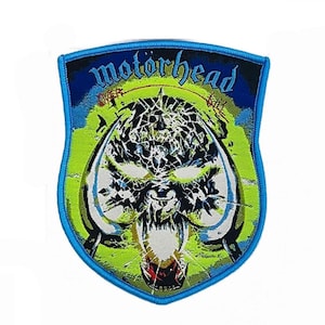 May include: A shield-shaped embroidered patch featuring the text motorhead in blue at the top. The design displays a stylised, fragmented skull graphic in black and white against a vibrant lime green background, all bordered by a bright blue edge.