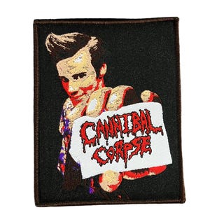 Peut inclure: Écusson rectangulaire avec un fond noir et une bordure marron. Il présente un graphisme d'une personne tenant une pancarte avec les mots "CANNIBAL CORPSE" en lettres rouges dégoulinantes.