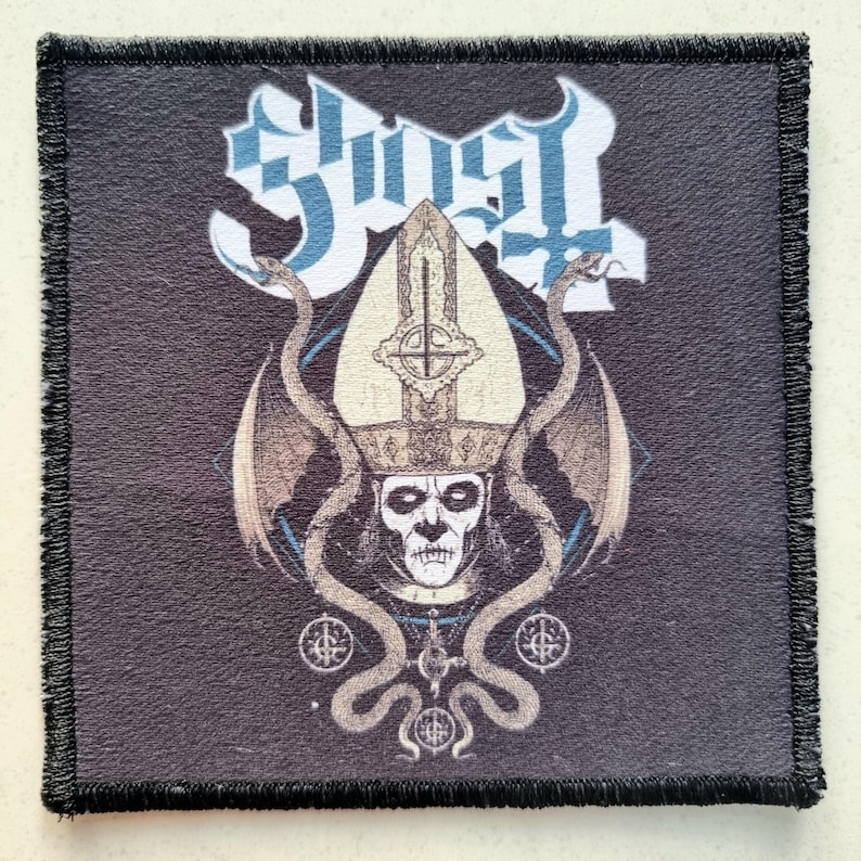 May include: Square black patch with a detailed graphic. The design features the word "Ghost" in blue and white, a skull wearing a papal mitre, and two snakes. The patch has a black stitched border.