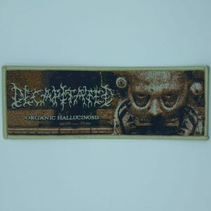 May include: A rectangular patch with the word "DECAPITATED" in a gothic font above the text "ORGANIC HALLUCINOSIS". The patch features a sepia-toned image of a face with a distorted appearance. The border is a light olive green.