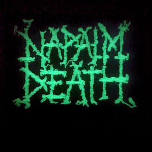 May include: A green glow-in-the-dark patch with the text "NAPALM DEATH" in a stylized font.