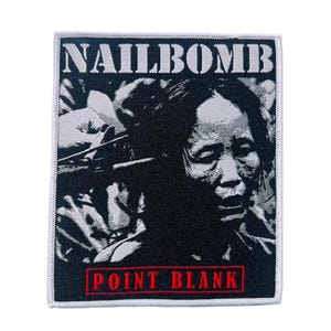 Pode incluir: Patch quadrado com uma imagem a preto e branco de um rosto. O texto "NAILBOMB" está no topo e "POINT BLANK" a vermelho na parte inferior. O patch tem uma borda branca.