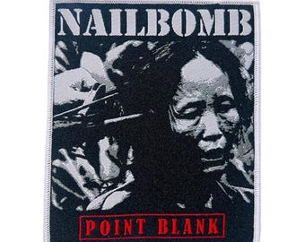 May include: A square patch featuring a black and white portrait of a person. The text "NAILBOMB" is at the top, with "POINT BLANK" in red at the bottom. The patch is edged with a white border.