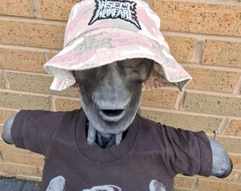 May include: A mannequin wearing a pink and grey camouflage bucket hat with the text "INSECT WARFARE" and a brown t-shirt with a graphic print. The mannequin is grey and has a neutral expression.