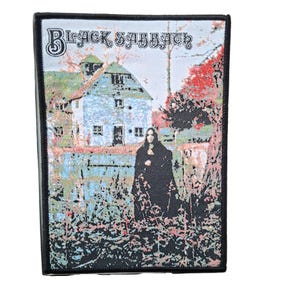 Puede incluir: Parche rectangular con el texto "Black Sabbath" sobre una imagen de una casa y una figura con una capa negra. La imagen tiene un borde negro y está sobre un fondo blanco. La casa es azul claro y verde, con follaje rojo y verde.