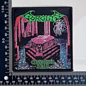 Puede incluir: Parche cuadrado negro con el nombre de la banda "Gorguts" en verde sobre una ilustración rosa y negra. La ilustración representa un sarcófago y el texto "Considered Dead" en verde. El parche mide aproximadamente 10 cm por 10 cm.