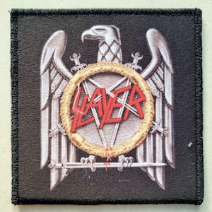 Puede incluir: Parche cuadrado negro con un águila plateada con las alas extendidas, un anillo dorado con un diseño de estrella y la palabra roja "SLAYER". El parche tiene un borde negro y es probablemente un emblema bordado.