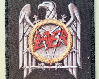 May include: Square black patch with a silver eagle emblem, a gold ring with a star design, and the red word "SLAYER". The patch has a black border and is likely an embroidered emblem.
