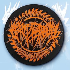 May include: A round, black patch with orange text and graphics. The text reads "SLAM-M-ING DEATHWARRIOR BRUTAL-DEATHGRIND" in a stylized, jagged font. The design features a circular saw blade motif.