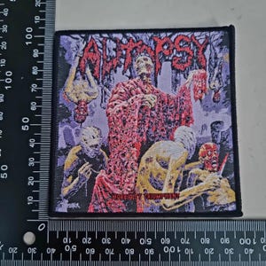 May include: Square patch with a macabre scene featuring the word "Autopsy" in red, stylized lettering. The patch depicts figures in a gory setting with a skeleton-like figure holding a severed head. The text "Rigor Mortis" is also visible.
