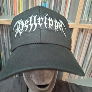 May include: Black baseball cap featuring the white embroidered text "Hellripper". The cap has a curved peak and a structured design. A collection of vinyl records is visible in the background.