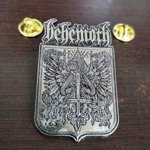 Puede incluir: Pin metálico plateado con la palabra "Behemoth" sobre un emblema detallado. El emblema presenta una cruz central, serpientes aladas y calaveras. También se ven dos cierres dorados.