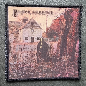 May include: Square patch with the Black Sabbath album art. The image shows a figure in a black cloak near a building and trees. The band's name is printed at the top in a stylized font. The patch has a black border.
