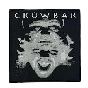 May include: A square black fabric patch featuring the white text CROWBAR at the top. Below the text is a high-contrast, stylized graphic of a face in shades of grey and white, depicted with a distorted, layered appearance and an open mouth.