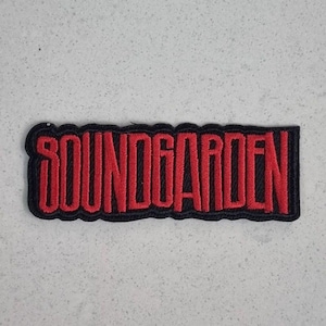 May include: A rectangular patch with the word "SOUNDGARDEN" embroidered in red thread. The text is outlined with a black border, creating a bold contrast. The patch is likely for clothing or accessories.