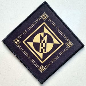 Op de afbeelding: Een zwart en goud Machine Head-patch. De vierkante patch heeft een zwarte rand en de naam van de band in goud. Het centrale ontwerp bevat een gouden ruit, een gouden cirkel en de letters "MH".