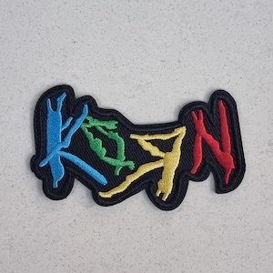 May include: Embroidered patch with the letters "KDN" in blue, green, yellow, and red. The letters are stitched onto a black background with a black border. The patch is on a light gray surface.
