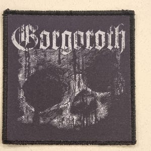 May include: A square, black patch featuring a white skull design and the word "Borgoroth" in a gothic font. The patch has a black border and a textured surface.
