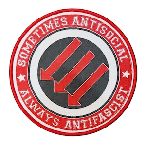 May include: Circular patch with red and white edging, displaying the text "SOMETIMES ANTISOCIAL ALWAYS ANTIFASCIST." The centre has a black circle with three red downward-pointing arrows. Two white stars are on the patch.