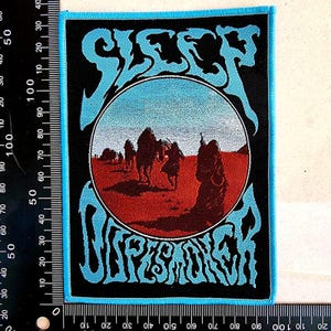 Peut inclure: Un écusson rectangulaire avec un fond noir et une bordure bleu clair. L'écusson présente les mots "SLEEP" et "DOPESMOKER" en lettres stylisées bleu clair. Une image circulaire au centre représente une scène de désert avec des personnages et des chameaux.