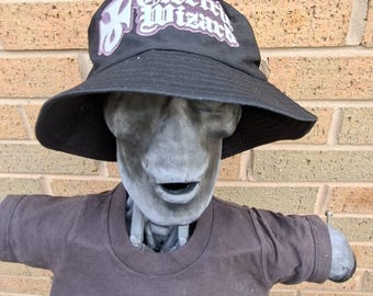 May include: A black bucket hat with the text "Electric Wizard" in white and purple lettering. The hat is worn by a grey statue wearing a brown t-shirt with a graphic print.