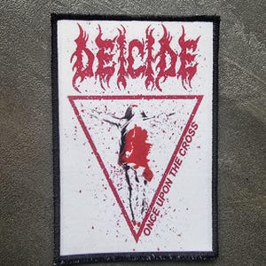 May include: A rectangular patch with a black border. The patch features the word "DEICIDE" in red, stylized lettering above a red triangle. Inside the triangle is a red and black illustration of a figure with outstretched arms. The text "ONCE UPON THE CROSS" is printed along the triangle's edge.