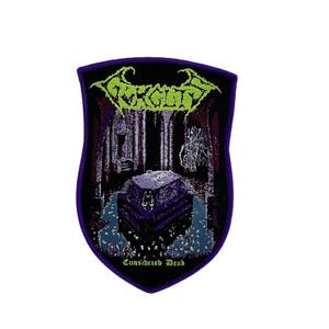 Könnte beinhalten: Ein schildförmiger, bestickter Aufnäher mit violettem Rand. Das Design zeigt ein dunkles, gotisches Interieur mit einem zentralen Steinsarkophag, einer geisterhaften Figur sowie dem Text "Carcass" in Limettengrün oben und "Considered Dead" unten.