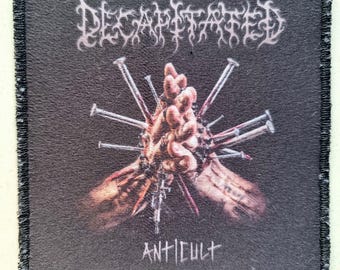 May include: A black square patch with the word "DECAPITATED" in white, stylized lettering. The patch features a graphic of hands pierced with nails, with the word "ANTICULT" below.