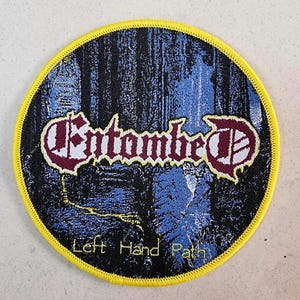 Puede incluir: Un parche bordado negro y amarillo con el nombre de la banda "Entombed" en letras blancas y rojas. El texto "Left Hand Path" está impreso en blanco debajo del nombre de la banda.